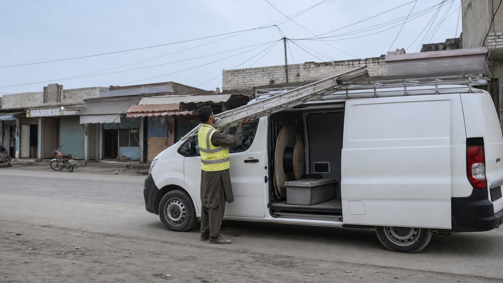 A technician in a hi-vis vest lifting a ladder from the roof of a white service van parked on a quiet high street, with a cable drum inside the open side door